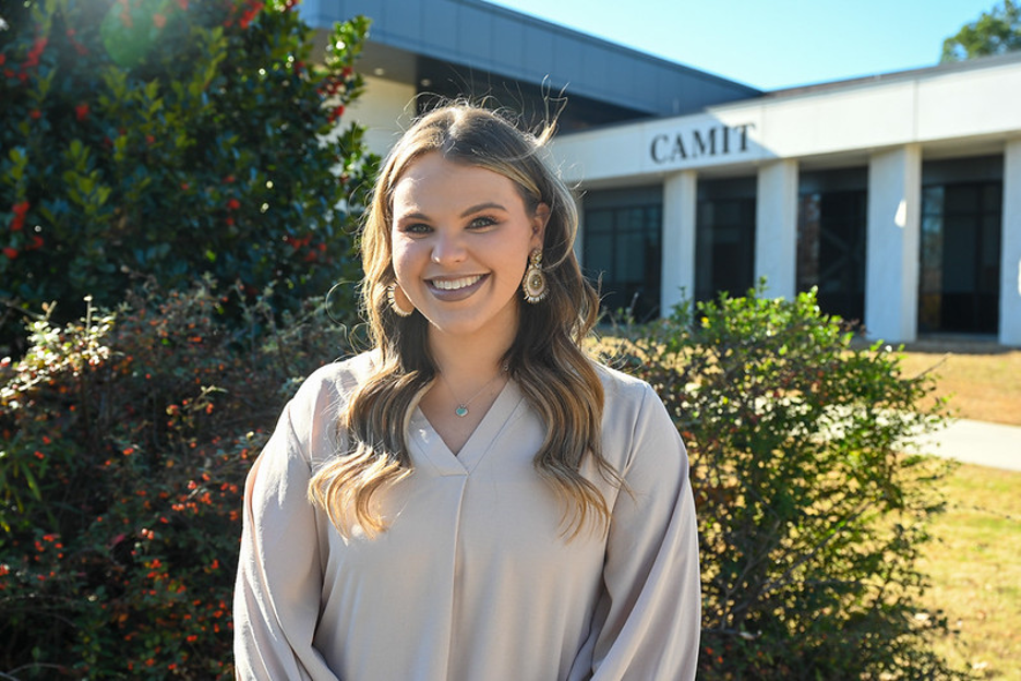 A woman stands outside a building, smiling at the camera. She is wearing a light tan long-sleeve top with small decorative dots. Behind her are large windows, a light-colored wall, and some landscaping with plants and stones.