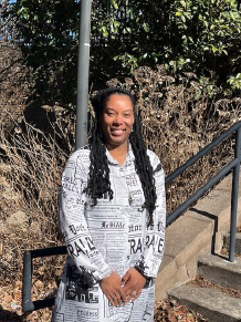 A person standing outdoors near a staircase and metal handrail, smiling at the camera. They are wearing a black-and-white patterned dress and have long braided hair, with shrubs and a campus-style walkway visible in the background.