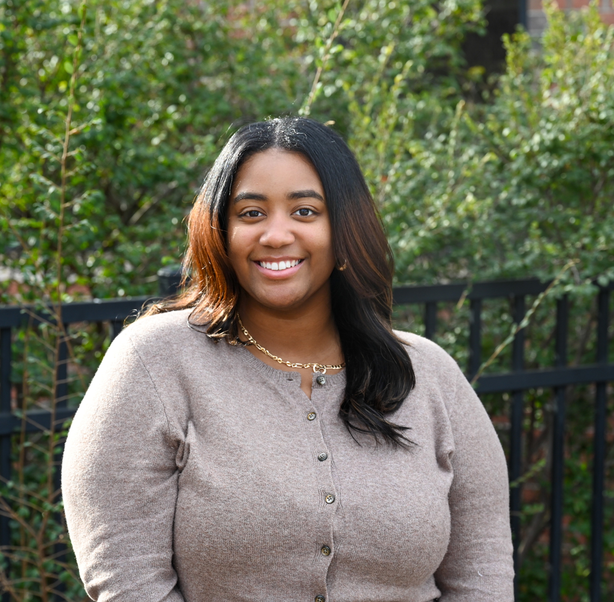 A smiling individual stands outdoors in front of greenery and a black metal fence. They are wearing a light beige button-front sweater and a gold necklace, with sunlight illuminating their face and hair. The background features softly blurred trees and shrubs, creating a natural campus-like setting.
