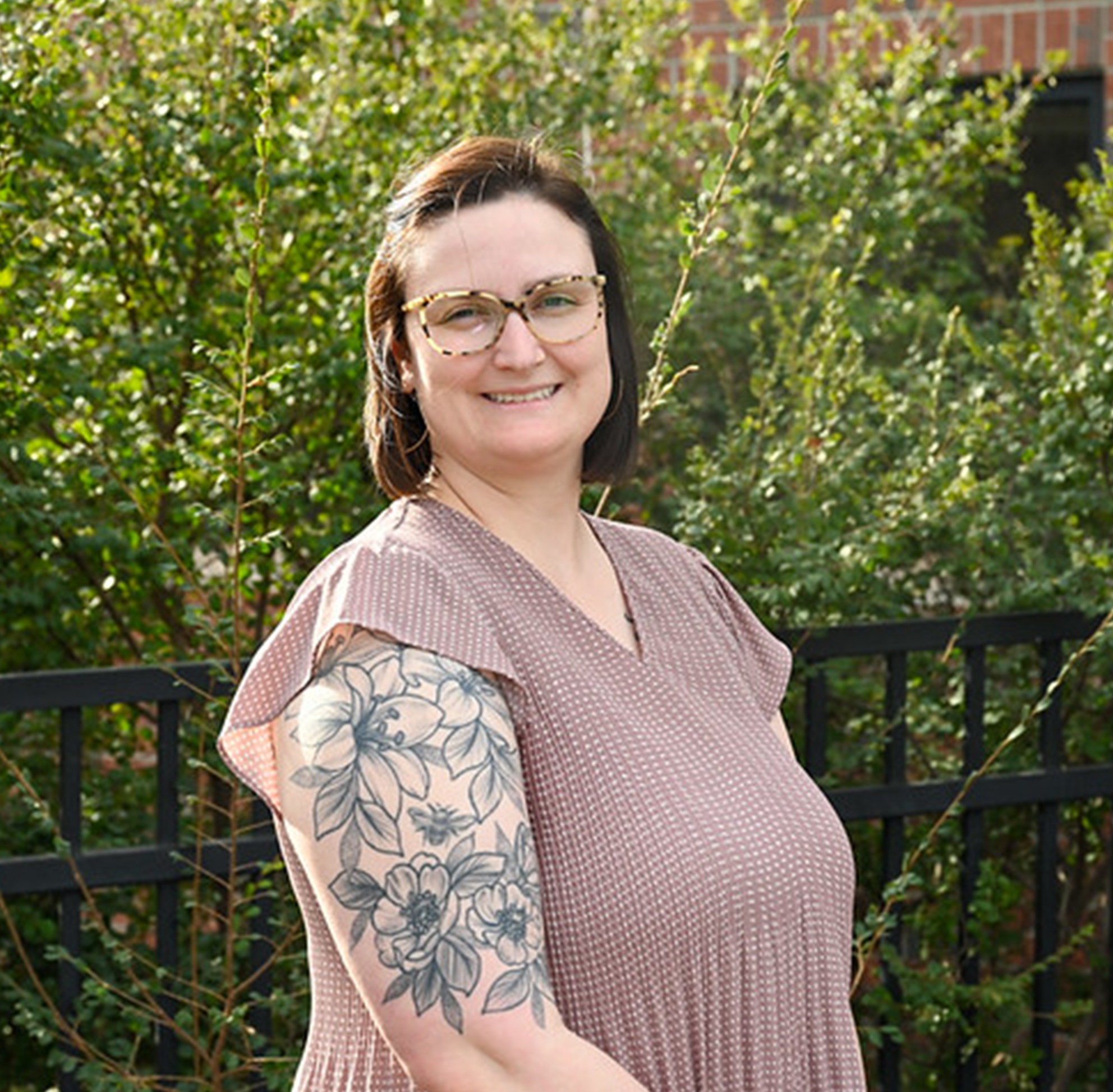 A smiling woman with shoulder-length dark hair and patterned glasses poses outdoors. She is wearing a mauve, short-sleeved, patterned top that reveals a large floral tattoo on her right upper arm. The background features green bushes and a black metal fence in front of a brick building.