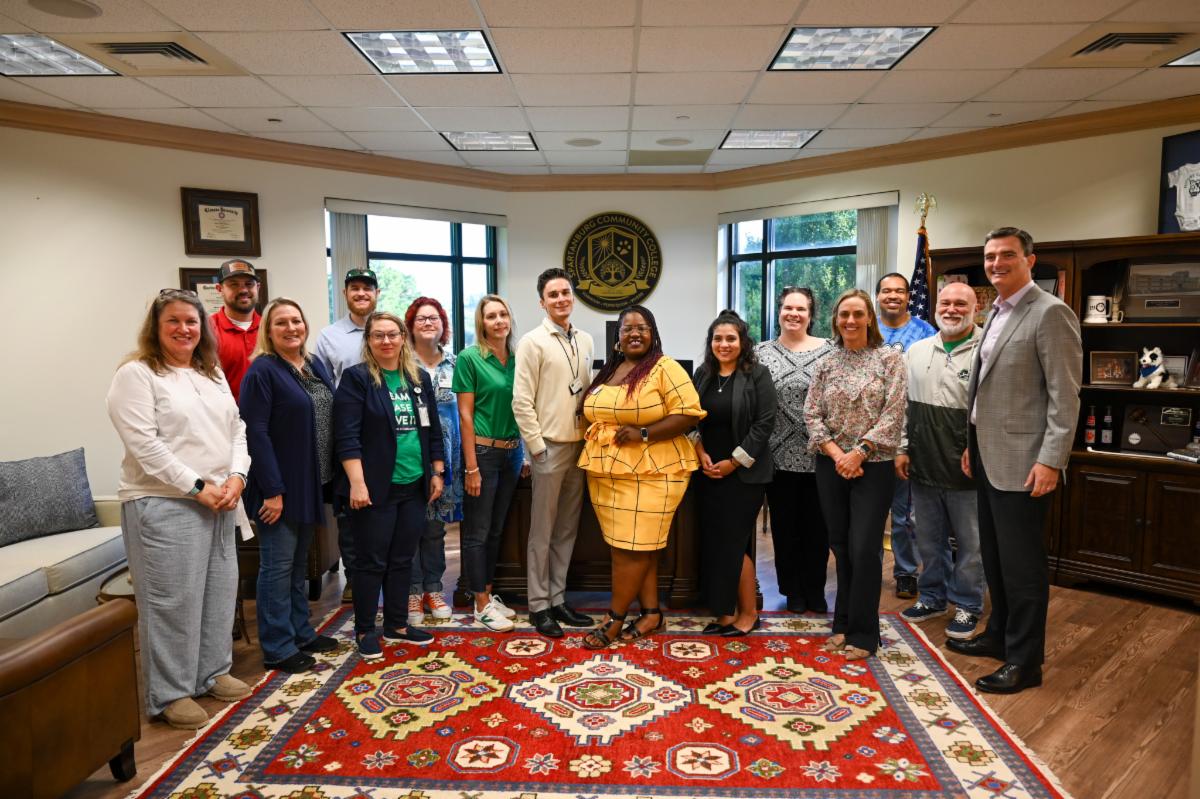 A group of sixteen professionally dressed adults pose for a group photo inside an office at Spartanburg Community College. The individuals stand side-by-side on a large, intricately patterned red oriental rug, positioned in front of a wooden desk and a circular college seal mounted on the wall. Large vertical windows let in natural light, while wooden furniture, and diplomas
