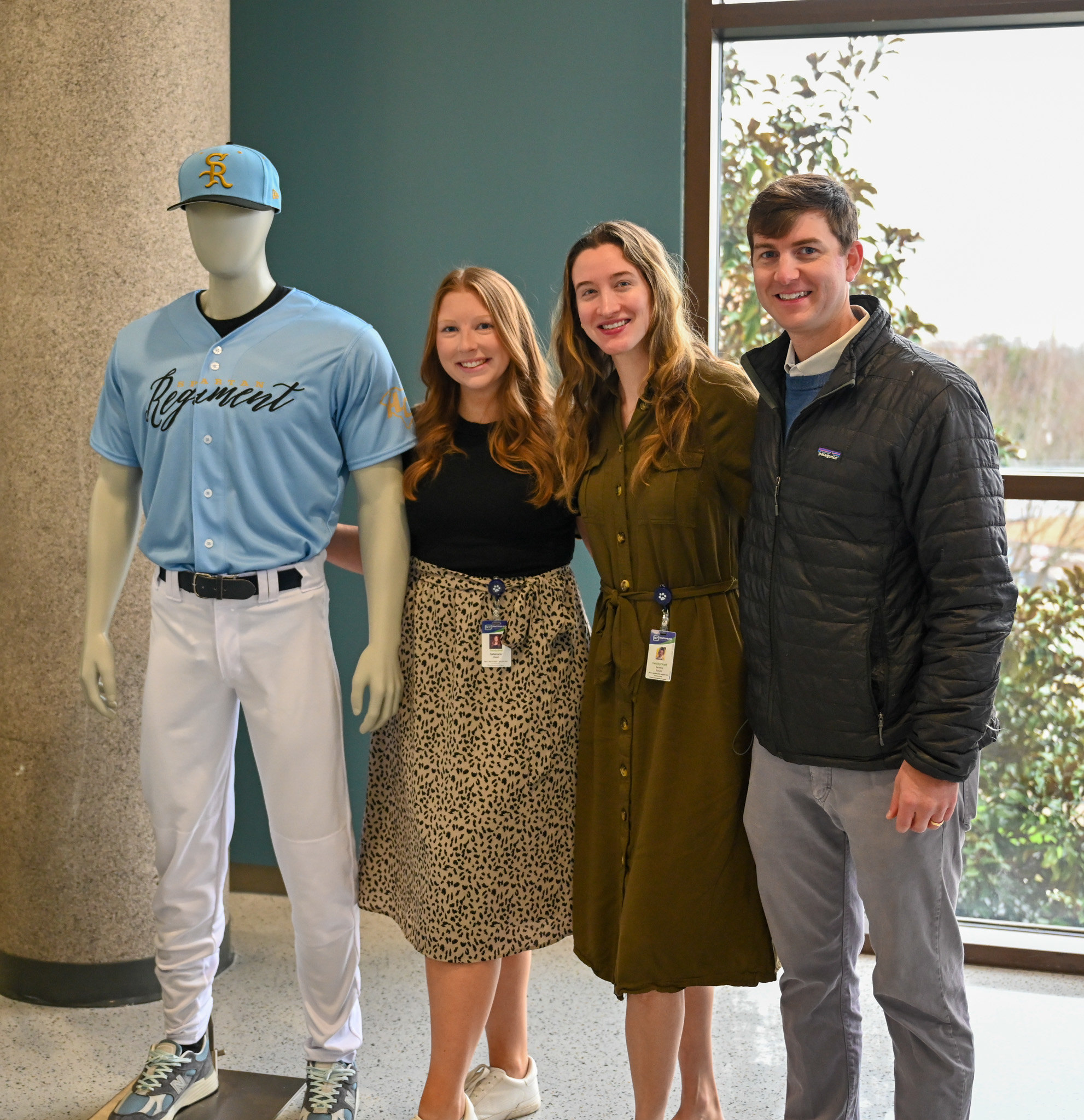 This image is a portrait of three people standing next to a mannequin displaying a baseball uniform by a large window with trees visible outside.