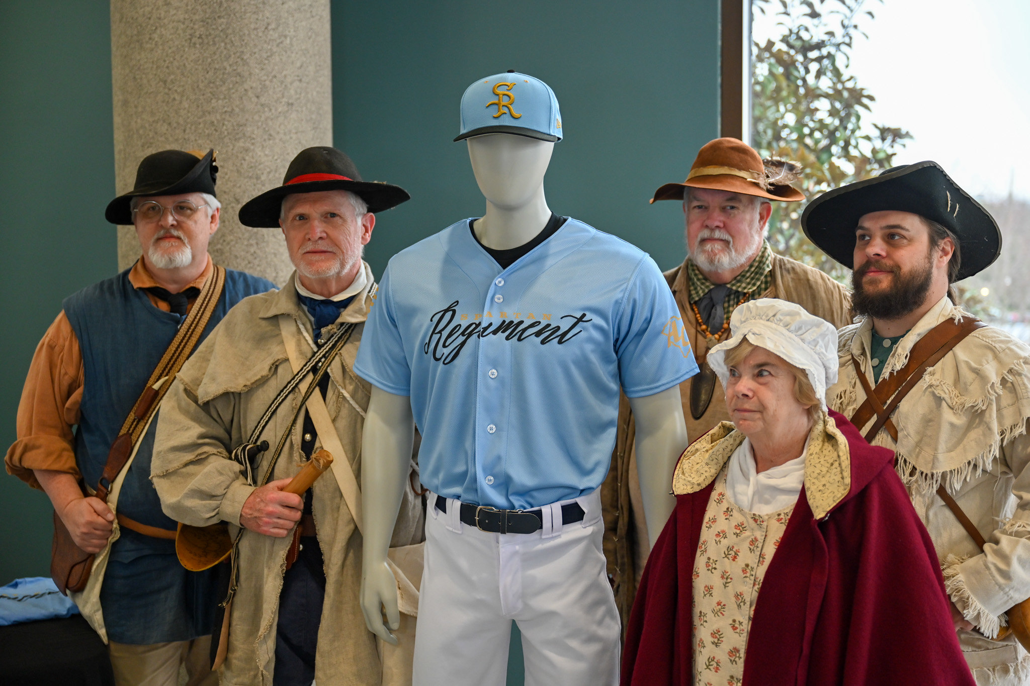 A mannequin wearing a light blue Spartan Regiment baseball uniform stands surrounded by people dressed in colonial-era reenactment clothing inside a building.