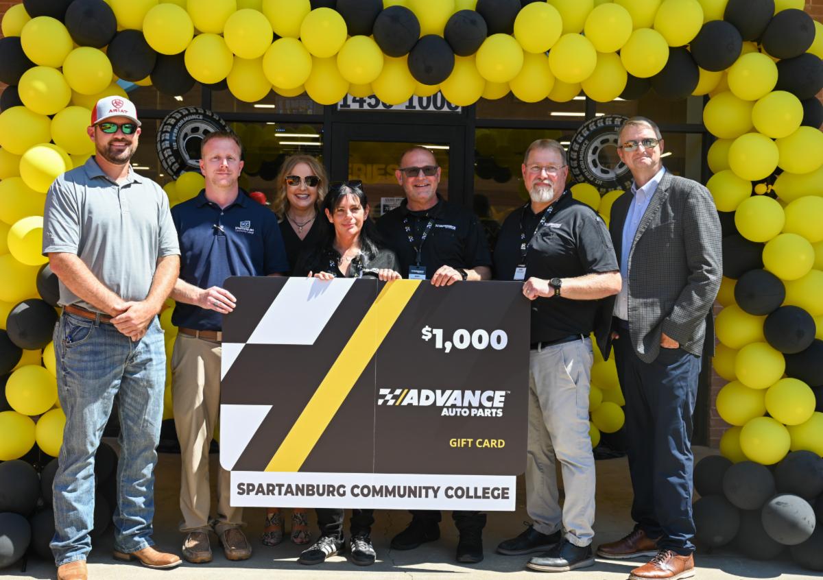 Seven people stand in front of an Advance Auto Parts store entrance decorated with a large black-and-yellow balloon arch. The group poses behind an oversized ceremonial $1,000 Advance Auto Parts gift card labeled for Spartanburg Community College, celebrating a partnership, donation, or community event. Bright sunlight illuminates the scene, with the store entrance and decorative tire displays