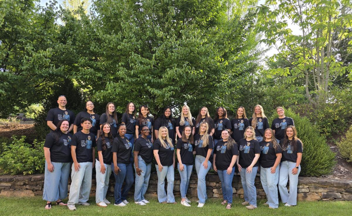 A group of approximately 23 students and faculty members pose outdoors in front of lush green trees and landscaping. The group is arranged in two rows, wearing matching black T-shirts and light blue jeans, standing on a grassy lawn beside a stone retaining wall. The setting appears to be a campus or garden environment, with bright natural sunlight and a celebratory atmosphere.