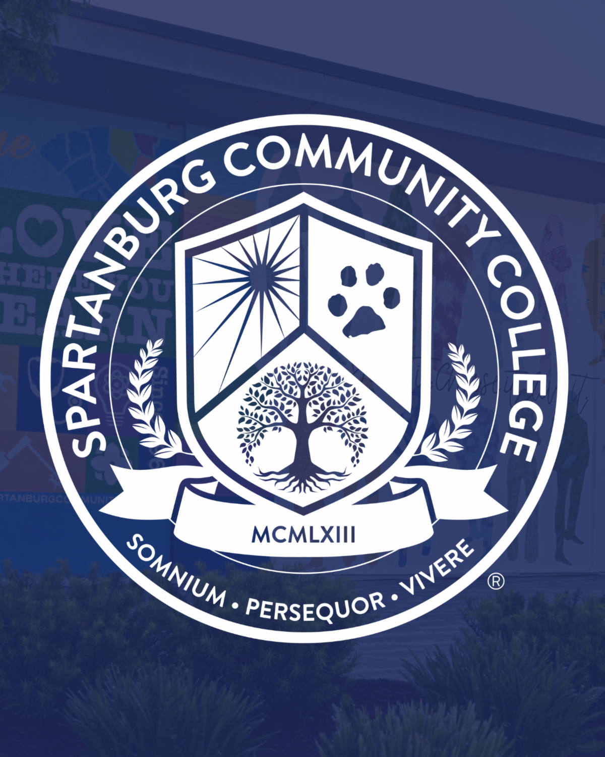 The official seal of Spartanburg Community College features a dark blue circular emblem with white text and graphics. The college name curves around the top border. Inside the center is a shield divided into three sections: a stylized rising sun, an animal paw print, and a detailed tree with deep roots. Below the shield is a banner displaying the Roman numerals 