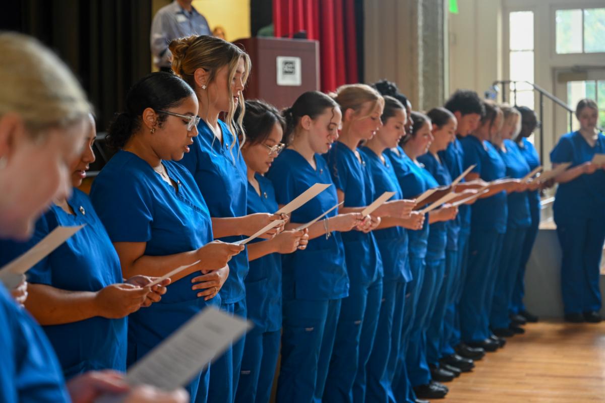 A side-angle, eye-level shot captures a long row of diverse nursing students standing side-by-side on a wooden floor in a brightly lit auditorium during a professional pinning or oath-taking ceremony. All individuals are wearing identical royal blue scrubs, holding white papers or printed pledges in their hands, and looking down to read along.