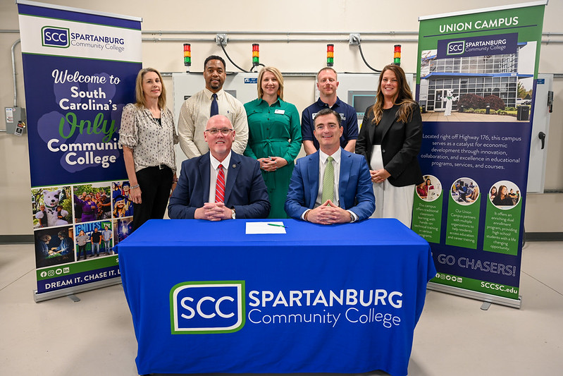 Seven staff and community leaders pose behind and in front of a table with Spartanburg Community College branding during a partnership signing event inside a technical training facility, flanked by SCC promotional banners highlighting the Union Campus and South Carolina community college programs
