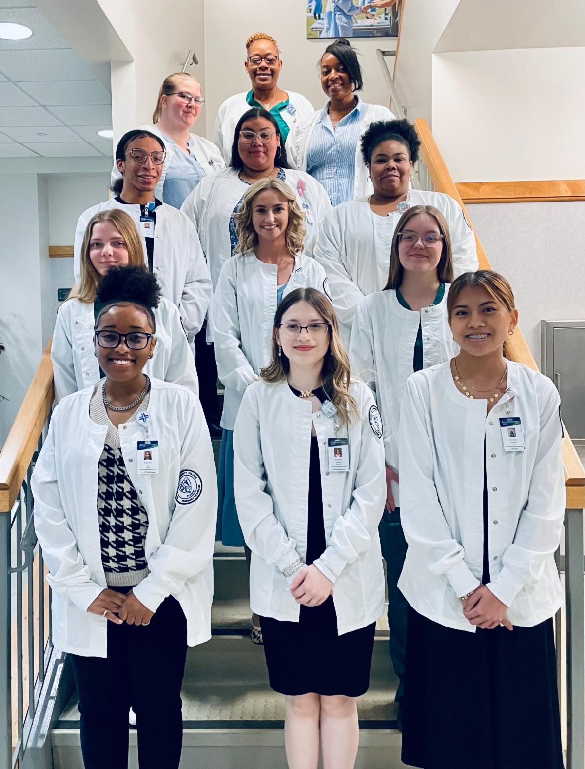 A group photo of ten diverse medical or nursing students posing on an indoor staircase. The students are arranged in tiers up the stairs, and all are wearing white laboratory coats or clinical jackets over their clothing or scrubs. Many have identification badges clipped to their lapels. The group includes individuals of various ethnicities and backgrounds, all smiling directly at the camera.