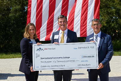 Three professionals standing outdoors in front of a large American flag, holding an oversized ceremonial check for $250,000.00 from USA Rare Earth presented to Spartanburg Community College for a community investment fund in Blacksburg, South Carolina, dated September 9, 2026.