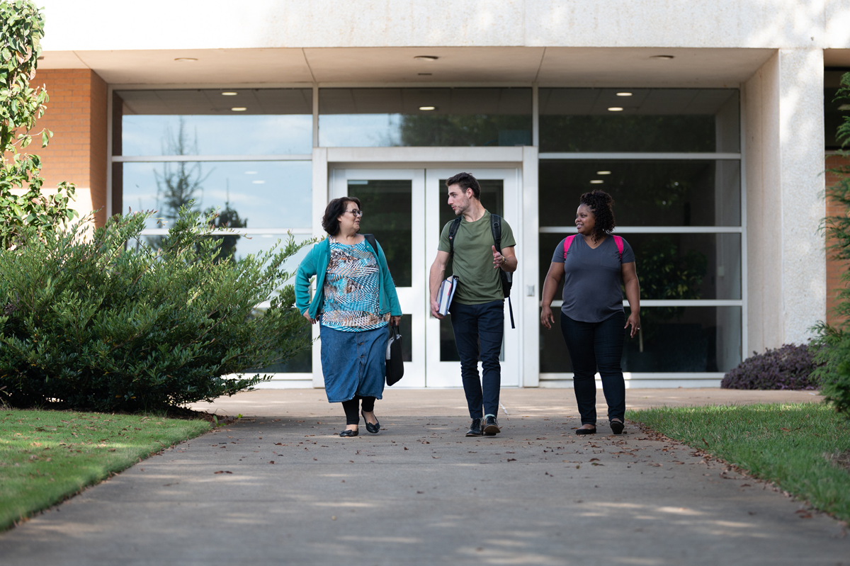 Three individuals walk side by side along a campus walkway in front of a building entrance with large glass doors and windows, carrying backpacks and folders, surrounded by trimmed bushes and greenery, with soft natural daylight creating a clean and professional college campus scene.