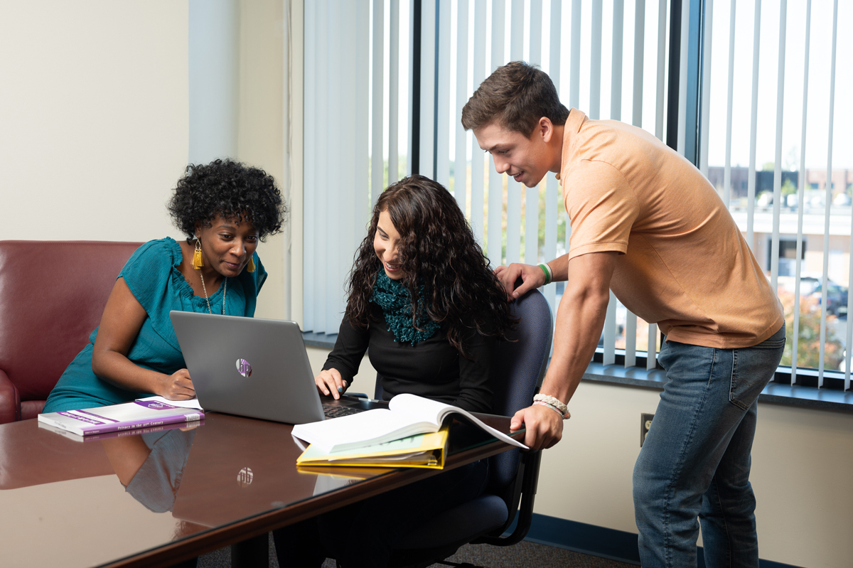 Three students collaborate around a laptop at a conference table inside a bright classroom or study space, with one seated individual typing while another leans over the shoulder and a third sits nearby with books and open binders, surrounded by vertical window blinds and natural daylight creating a focused academic environment.