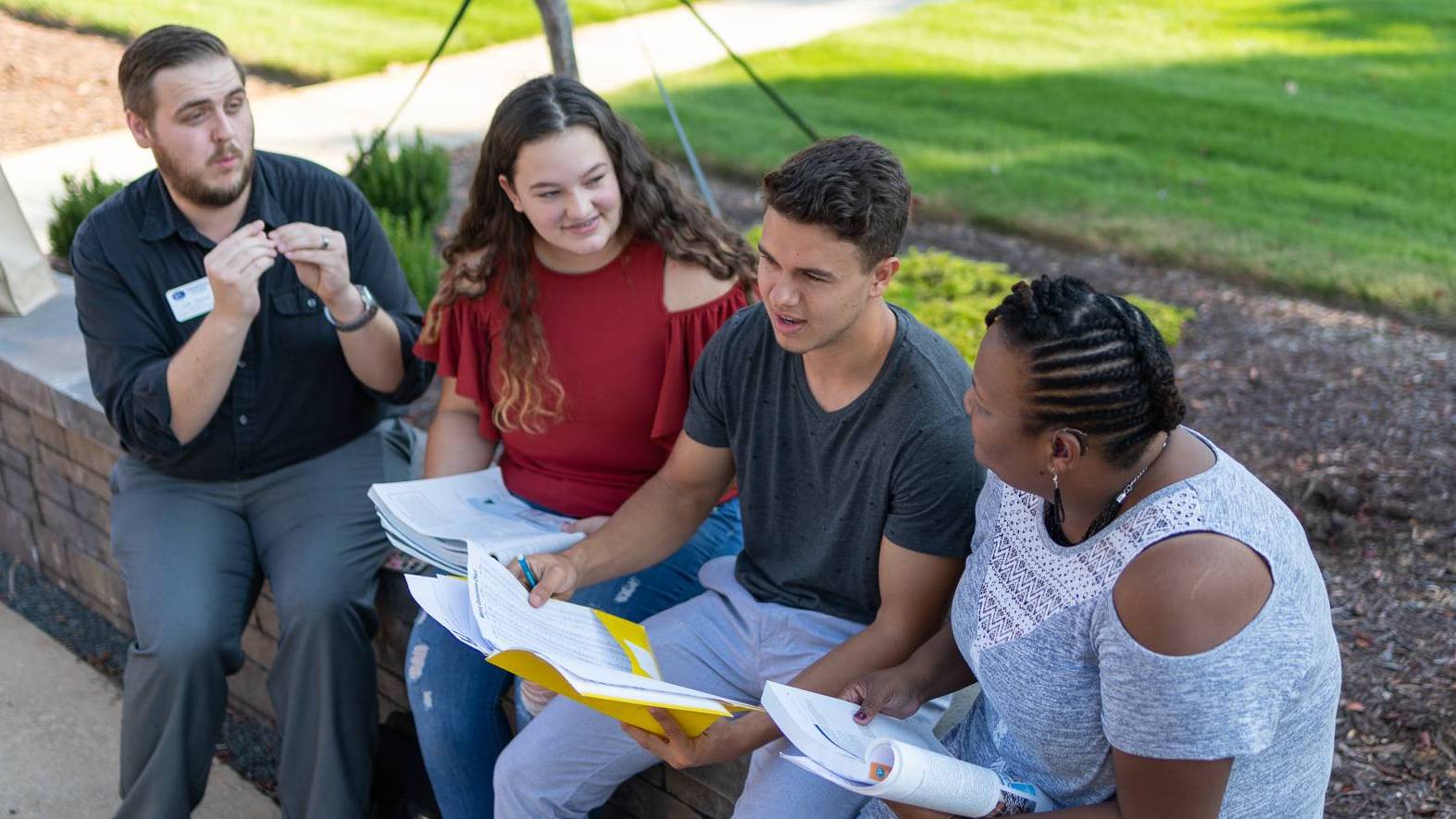 Group of four individuals seated outdoors on a low brick wall in a campus setting, collaborating over printed documents and folders, with one individual using hand gestures while communicating, and greenery, a walkway, and landscaped areas in the background under bright natural daylight.