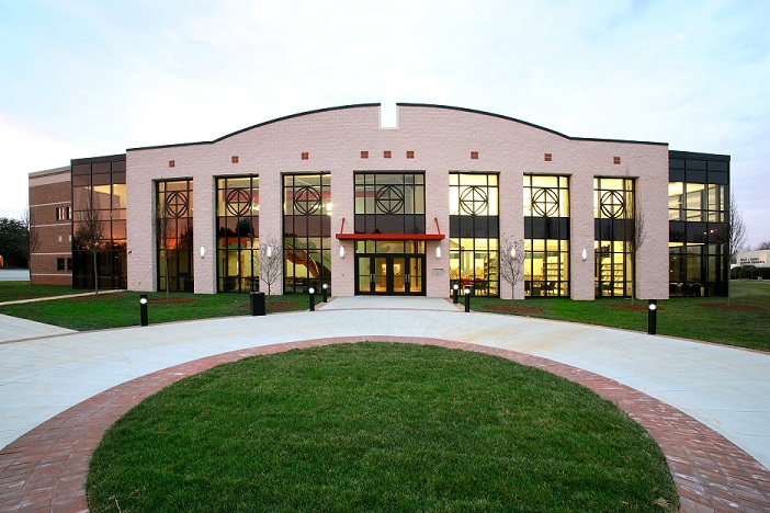 Exterior view of a modern campus building with a curved brick façade and large glass windows, featuring a central entrance with red awning, symmetrical window design, and well-maintained landscaping including green grass and a circular brick walkway in the foreground under soft daylight.