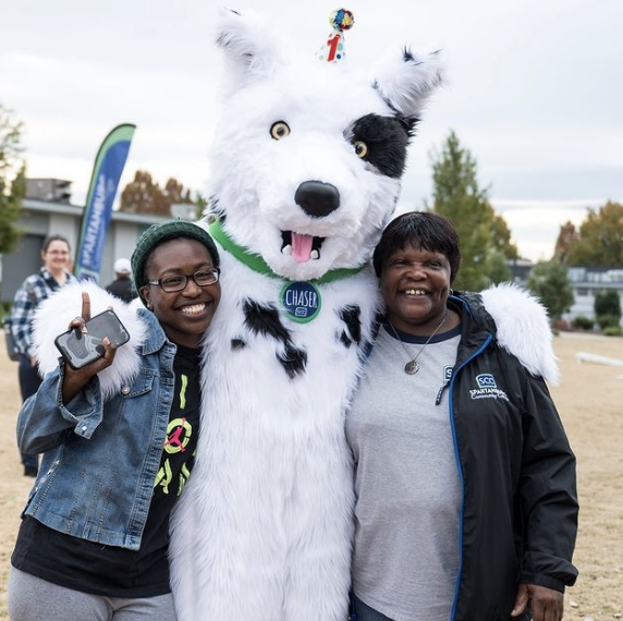 Outdoor campus event scene featuring Chaser, the Spartanburg Community College mascot, standing in the center wearing a green bandana and name tag, with two individuals on either side, one holding a phone, all standing on a grassy area with tents, banners, and trees in the background under soft daylight.