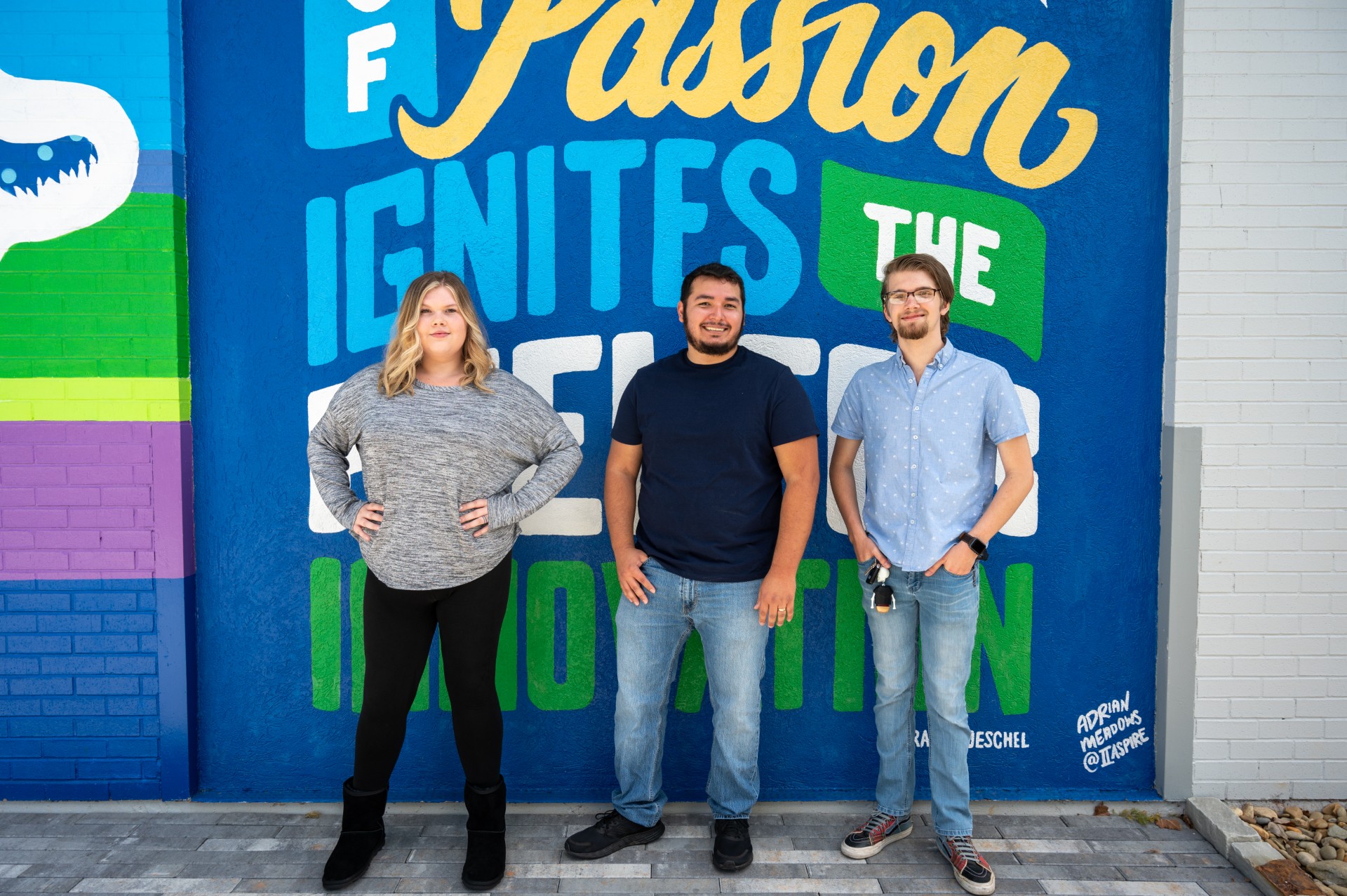 Three individuals stand side by side in front of a large, colorful mural wall featuring bold text reading “Fashion Ignites the Motion,” with bright blue, green, and yellow painted sections and graphic elements, wearing casual clothing and posing on a paved walkway in an outdoor urban or campus setting.